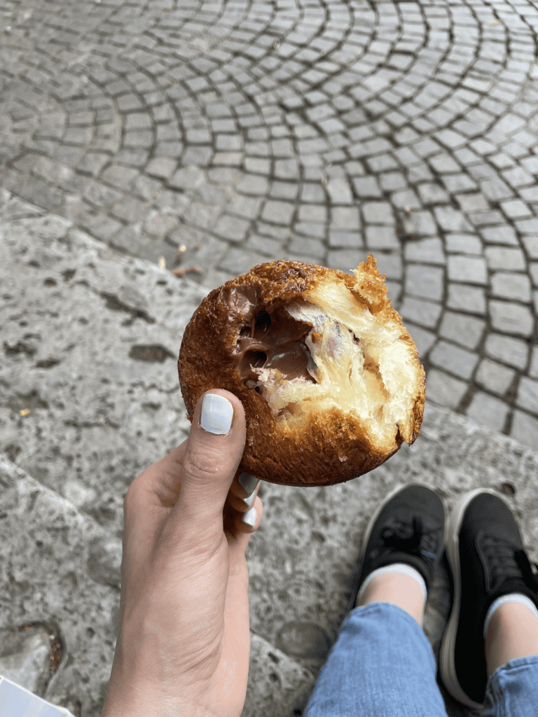 a hand holding a sphere croissant with a bite taken out of it in a park