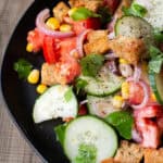 a close-up of a panzanella on a black plate made of bread, cucumbers, basil, corn, tomatoes, olives, and shallot