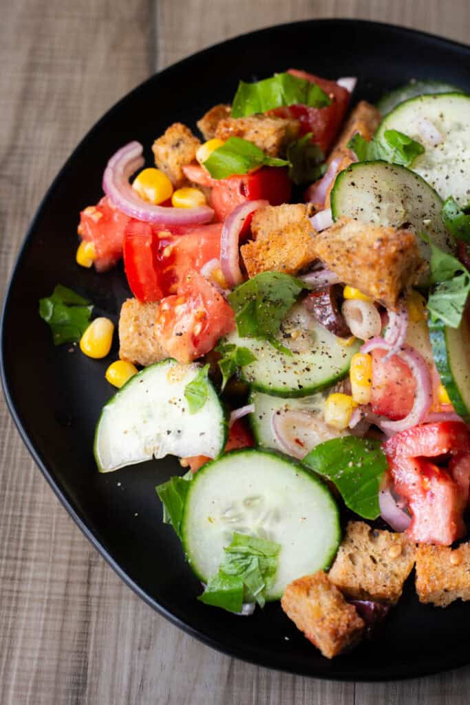 a close-up of a panzanella on a black plate made of bread, cucumbers, basil, corn, tomatoes, olives, and shallot